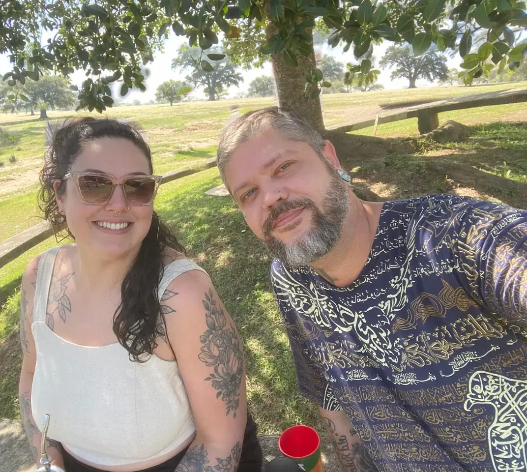 Amanda, de óculos escuros e regata clara, com tatuagens florais nos braços, e André, de barba grisalha e camisa azul-marinho estampada, sentados lado a lado à sombra de uma árvore, num banco de praça